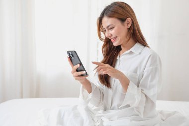 Beyaz gecelikli bir kadın hafta sonu uyanıp dinleniyor ve cep telefonuyla oynuyor ekmek yiyor ve beyaz yatak odasında çay içiyor. Sabah tatili.