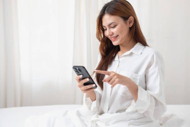 Beyaz gecelikli bir kadın hafta sonu uyanıp dinleniyor ve cep telefonuyla oynuyor ekmek yiyor ve beyaz yatak odasında çay içiyor. Sabah tatili.