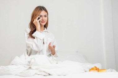 Beyaz gecelikli bir kadın hafta sonu uyanıp dinleniyor ve cep telefonuyla oynuyor ekmek yiyor ve beyaz yatak odasında çay içiyor. Sabah tatili.