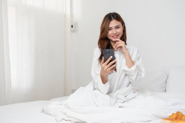 Beyaz gecelikli bir kadın hafta sonu uyanıp dinleniyor ve cep telefonuyla oynuyor ekmek yiyor ve beyaz yatak odasında çay içiyor. Sabah tatili.