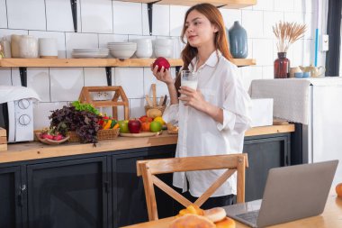 Beyaz gömlekli güzel yüzlü bir kadın mutfakta ekmek, sebze, meyve ve sütle sağlıklı bir kahvaltı hazırlıyor ve yemek dersleri için dizüstü bilgisayarını açıyor. sağlıklı yemek fikirleri.