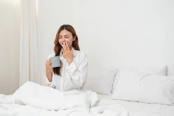 stock image Woman in white nightgown waking up on weekend morning resting and relaxing playing with laptop mobile phone Eating bread and drinking tea in glass inside white bedroom.  Morning vacation concept.