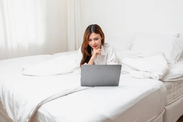 stock image Woman in white nightgown waking up on weekend morning resting and relaxing playing with laptop mobile phone Eating bread and drinking tea in glass inside white bedroom.  Morning vacation concept.