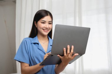 Güzel yüzlü, mavi tişörtlü, beyaz yatak odasındaki beyaz yatakta oturan, dizüstü bilgisayarlı ve tatilde akrabalarıyla konuşan genç Asyalı bir kadın. tatil kavramı.