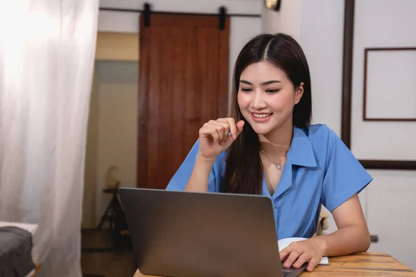 stock image Asian woman with a beautiful smile and face sits comfortably on her desk at home with a laptop and notebook for relaxing and relaxing in a bright morning. holiday vacation concept.