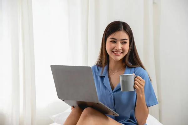 stock image Young Asian woman with beautiful face, long hair in blue shirt sitting on white bedroom bed at home with cup of coffee and video call with laptop talking to relatives on holiday. vacation concept.