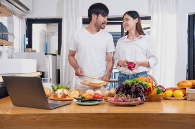 Meyve sebzelerle yemek yapan ve mutfakta dizüstü bilgisayar kullanan genç Asyalı çift aile içinde birlikte yemek pişiriyor. Aile kavramı..