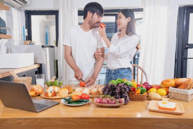 Meyve sebzelerle yemek yapan ve mutfakta dizüstü bilgisayar kullanan genç Asyalı çift aile içinde birlikte yemek pişiriyor. Aile kavramı..