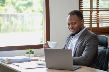 Siyah takım elbiseli Afro-Amerikan işadamı şirketin ofisteki laptopunu kontrol ettikten sonra rahatlamak için elinde bir fincan kahve tutuyor..