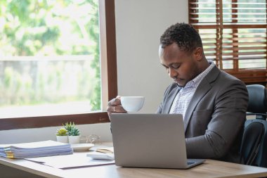 Siyah takım elbiseli Afro-Amerikan işadamı şirketin ofisteki laptopunu kontrol ettikten sonra rahatlamak için elinde bir fincan kahve tutuyor..