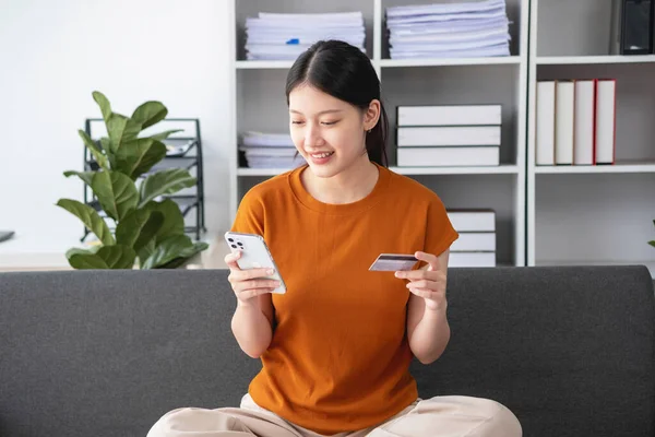 Young adult Asian female consumer holding credit card and smartphone sitting on the floor at home doing online banking transactions. E-commerce virtual shopping, secure mobile banking concept.