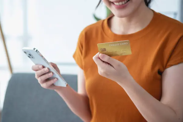 Young adult Asian female consumer holding credit card and smartphone sitting on the floor at home doing online banking transactions. E-commerce virtual shopping, secure mobile banking concept.