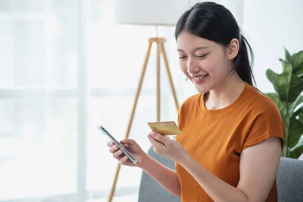 Young adult Asian female consumer holding credit card and smartphone sitting on the floor at home doing online banking transactions. E-commerce virtual shopping, secure mobile banking concept.