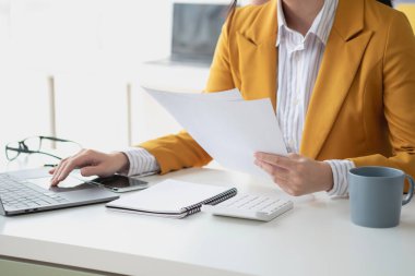 Sarı takım elbiseli Asyalı bir işkadını ya da yönetici şirket gelirini ve ofis içindeki masrafları hesaplamak için dizüstü bilgisayar ve hesap makinesi kullanıyor..