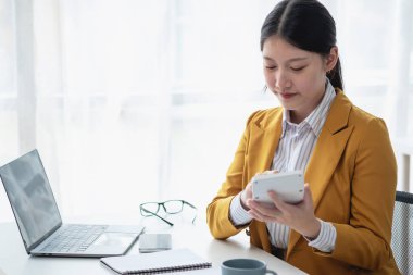 Sarı takım elbiseli Asyalı bir işkadını ya da yönetici şirket gelirini ve ofis içindeki masrafları hesaplamak için dizüstü bilgisayar ve hesap makinesi kullanıyor..