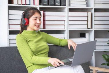 Yeşil uzun kollu gömlekli, güzel yüzlü Asyalı kadın oturmuş film seyrediyor dizüstü bilgisayar, cep telefonu ve kulaklıkla evde mutlu bir şekilde müzik dinliyor..