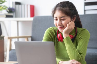 Yeşil uzun kollu gömlekli, güzel yüzlü Asyalı kadın oturmuş film seyrediyor dizüstü bilgisayar, cep telefonu ve kulaklıkla evde mutlu bir şekilde müzik dinliyor..