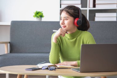 Yeşil uzun kollu gömlekli, güzel yüzlü Asyalı kadın oturmuş film seyrediyor dizüstü bilgisayar, cep telefonu ve kulaklıkla evde mutlu bir şekilde müzik dinliyor..