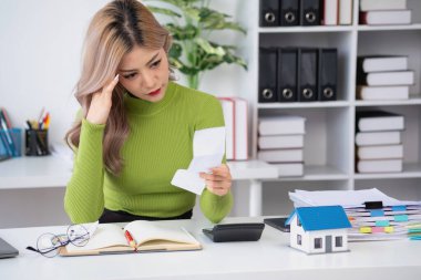 Asya 'lı bir kadın oturuyor ve hesaplarını yapıyor. Masasında hesap makinesi ve ev ile ipotek var. Ödemeler, ödenmeyen krediler, iş yerleri ve gayrimenkul konusunda endişeli.