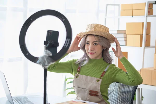 A cute Asian female blogger reviews products and camcorders on a home network, sells online, and presents fashion hats on social media. E-commerce business live stream vlogs new normal concept.