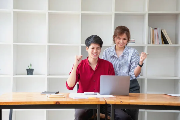 Cheerful business people using a laptop in an office. Happy young entrepreneurs smiling while working together in a modern workspace. Two young business people are sitting together at a table.