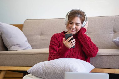 Kırmızı gündelik elbiseli güzel bir kadın evde kanepede dinlenirken müzik dinlemekten ve gülümsemekten zevk alıyor. Kulaklıklı genç bir kadın evde dizüstü bilgisayar ve akıllı telefon kullanıyor. Rahatla konsept.