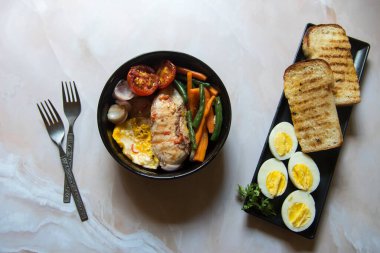 Ekmek dilimleri ve yumurtalarla servis edilen bir kasede soslu sebzeyle servis edilen ızgara tavuk filetosu. Üst görünüm, seçici odak.