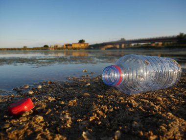 Yerde, arka planda bir kanal ve köprü bulunan boş bir plastik şişenin yakın plan çekimi..