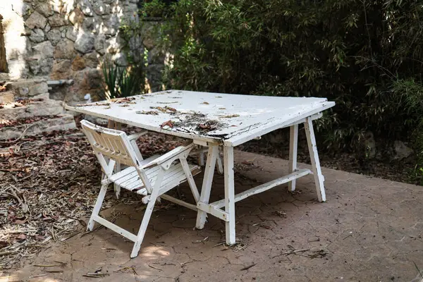 Sessiz bir açık havada, eski bir ahşap beyaz masa ve sandalye dağınık yaprakların arasında oturur..