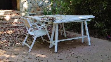 Sessiz bir açık havada, eski bir ahşap beyaz masa ve sandalye dağınık yaprakların arasında oturur..