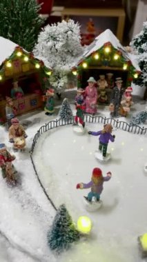Kapalı minyatür Noel oyuncağı, küçük insanlar bir buz pateni sahasında daireler çizerek kayıyor xmas caddesi pazarında çelenkle aydınlatılıyor, kışın Noel manzarası, 4k, dikey