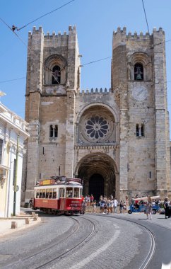 main facade of the cathedral of Lisbon, with the tramway number 28 passing through the door and tourists seeing the alfama district, vertical