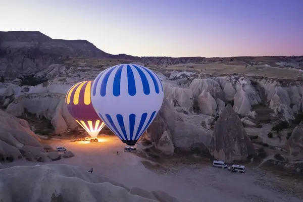 two hot air balloons full of tourists, about to take off at dawn between fairy chimneys, service vans around the balloons, cappadocia turkey, horizontal