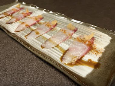 Bir tabak ince dilimlenmiş limonlu balık sashimi, bir parça sarımsak, paprika ve sos, kahverengi bir masada seramik tabakta deri paspas, tipik Japon yemeği, seçici odak noktası