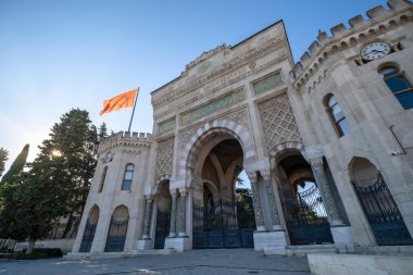 İstanbul Üniversitesi 'nin ana girişinde mavi gökyüzüne karşı sallanan hindi bayrağı, ağaçların arasından süzülen güneş ışınları, yatay