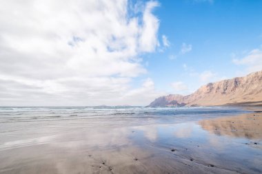 Arka planda kayalıklar bulunan Famara plajı kıyıya, Lanzarote Adası 'nın kuzeyine, Kanarya Adalarına, İspanya' ya, Caleta de Famara 'ya, yatay olarak yansımıştır.