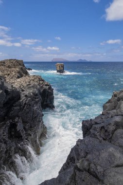 a cliff of black volcanic rock with a sea of turquoise water with waves lapping against the rocks, a picturesque rock rising out of the water, vertical clipart