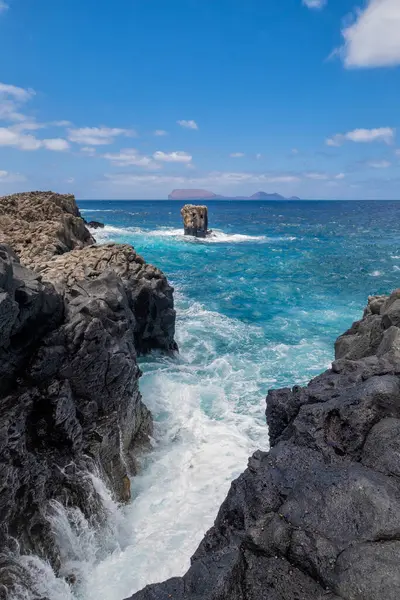 stock image a cliff of black volcanic rock with a sea of turquoise water with waves lapping against the rocks, a picturesque rock rising out of the water, vertical