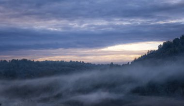 Misty morning landscape with soft pastel colors, dense fog covering hills, and a dramatic cloudy sky illuminated by the warm hues of sunrise, creating a serene and atmospheric view, panoramic clipart