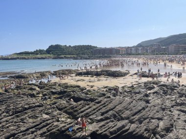 Castro Urdiales beach full of people on a summer day, with rocks in the foreground and front line apartments in the background, horizontal clipart