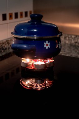 Çiçeklerle süslenmiş mavi bir güveç. Viteroseramik bir tabakta. Büyük bir elektrik kesintisi sırasında elektriksizliğin acil durum çözümü olarak mumların üzerinde ısıtılıyor.