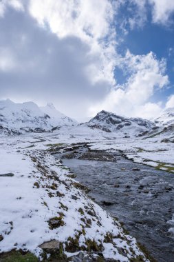 Karlı dağ manzarası, bulutlu bir gökyüzünün altındaki vadiden akan berrak bir dere, huzurlu bir dağda eriyen kışın manzarası ve doğal güzellik.