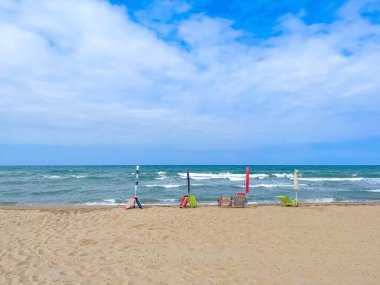 Sahilin ilk satırında yer ayırtmak için kumsala yerleştirilmiş dört kapalı plaj şemsiyesi ve plaj sandalyesi, kapalı şemsiyeler ve kumlu bir sahilde denize bakan boş plaj sandalyeleri.