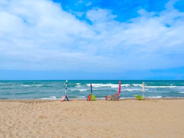 Sahilin ilk satırında yer ayırtmak için kumsala yerleştirilmiş dört kapalı plaj şemsiyesi ve plaj sandalyesi, kapalı şemsiyeler ve kumlu bir sahilde denize bakan boş plaj sandalyeleri.