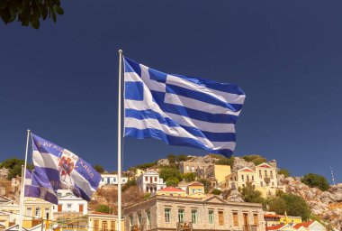 Yunan ulusal bayrağı, dağların mavi gökyüzünün arka planına ve Symi köyündeki eski evlerin cephelerine karşı. Yunanistan.