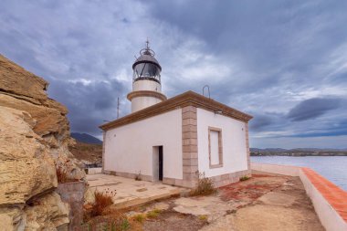 Cala Nans deniz feneri bulutlu gökyüzünün altındaki kayaların üzerinde, arka planda fırtınalı deniz, İspanya, Katalonya