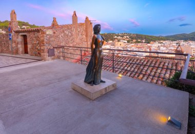 Tossa de Mar, Spain - September 14, 2014: The statue of Ava Gardner towers over the rooftops of Tossa de Mar at sunrise. The bronze sculpture is located just outside the historic walls of Vila Vella, with picturesque views of the town and surrounding clipart