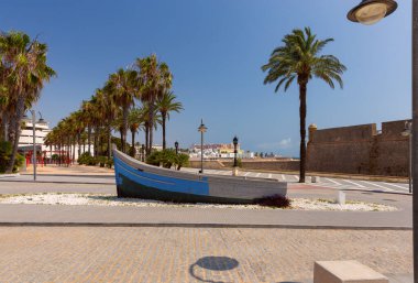 Açık mavi gökyüzünün altında dekoratif mavi tekne heykeli, palmiye ağaçları ve tarihi şehir mimarisinin yer aldığı Cadiz 'deki gezinti alanının manzarası, İspanya
