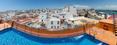 İspanya, Cadiz 'deki bir çatı havuzundan panoramik bir manzara, tarihi şehirlere tepeden bakan beyazlatılmış binalar, renkli çatılar, ve güneşli mavi bir gökyüzünün altındaki rıhtım.