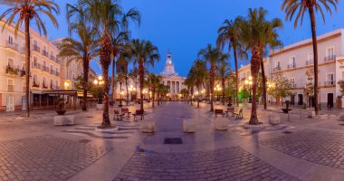 İspanya 'nın Cadiz kentindeki Plaza de San Juan de Dios meydanında, şafak vakti, aydınlık fıskiyeler, palmiye ağaçları ve tarihi binalar bulunmaktadır.
